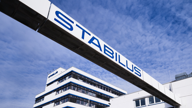 Firmengebäude der Stabilus Gruppe mit modernem Architekturdesign und einer markanten Brücke mit dem Schriftzug „STABILUS“ vor blauem Himmel.