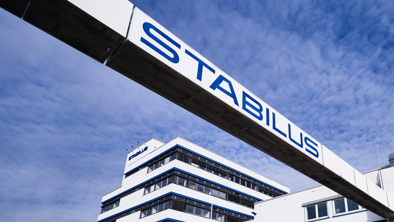 Firmengebäude der Stabilus Gruppe mit modernem Architekturdesign und einer markanten Brücke mit dem Schriftzug „STABILUS“ vor blauem Himmel.